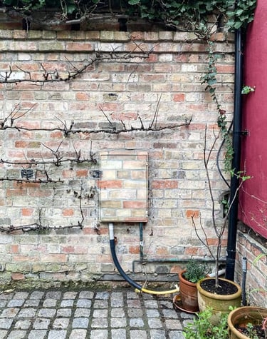 An electricity box attached to a garden brick wall painted in camouflage brick pattern.