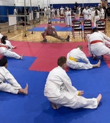 Martial arts students in white gi uniforms stretch on blue and red mats during a karate tournament.