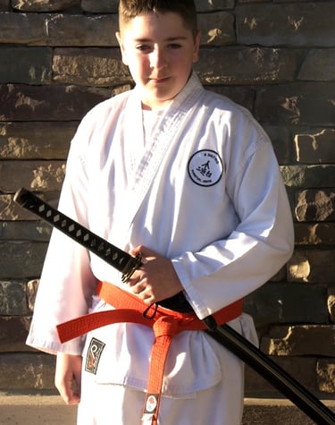 Young boy in a white martial arts gi and orange belt holding a katana sword in front of a stone wall.