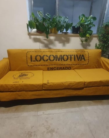 Sofá industrial de lona amarela com a marca Locomotiva Encerado e plantas em vasos ao fundo.