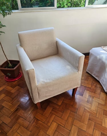Poltrona estofada bege com pernas de madeira em um piso de parquet em uma sala de estar iluminada.