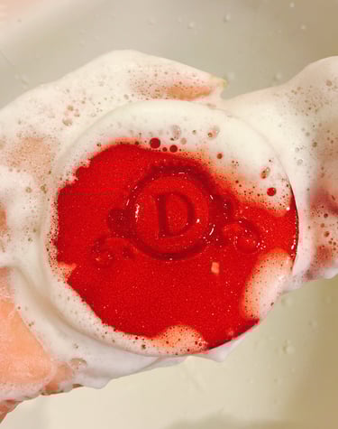 A round red bar of soap with a letter D engraving held in a hand covered in white soapy bubbles.