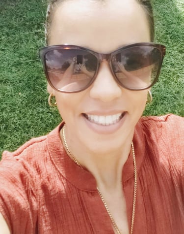 Smiling woman wearing tortoise shell sunglasses and gold jewelry in a green garden.