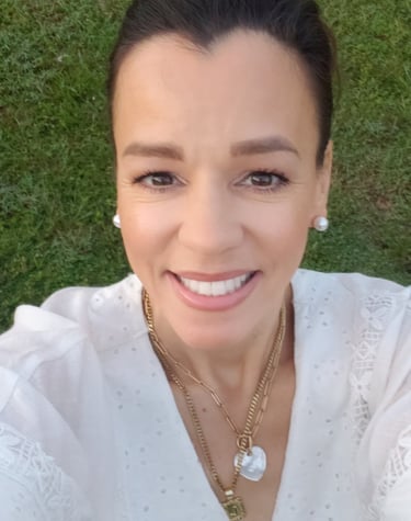 Smiling woman in white lace dress and gold necklaces outdoors against green grass.