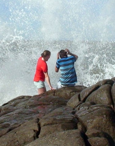 two backpackers on a rock being hit by a wave