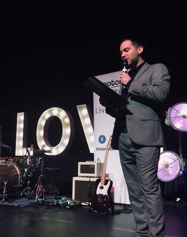 Alex hosting an awards function in a grey suit holding a clipboard