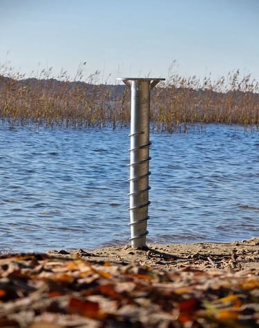 Galvanized steel ground screw anchor installed in sandy shore for lakeside dock foundation.