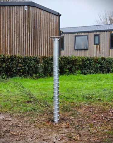 Galvanized steel ground screw foundation installed in a grassy backyard near wooden garden buildings.