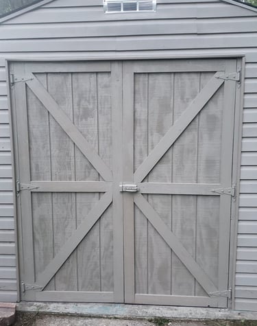 Gray wooden storage shed with double barn doors and cross-braces for backyard organization.
