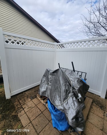 Backyard patio equipment protected by a heavy-duty silver tarp inside a white vinyl privacy fence.