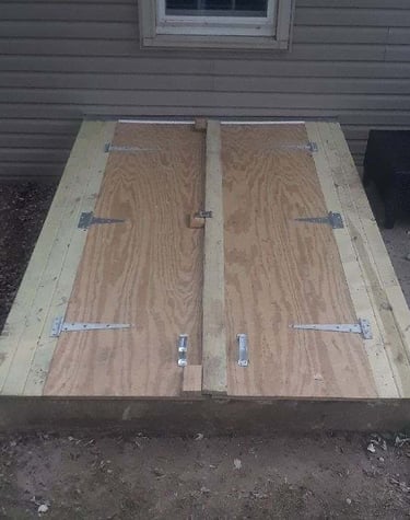 Custom-built wooden storm cellar doors made of plywood and lumber with metal hinges outside a home.