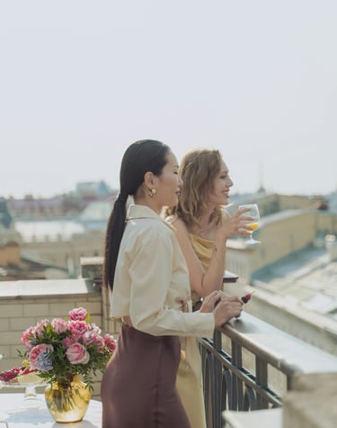 Woman with pink hair working remotely on a laptop in a modern hotel room with mountain views, next to a purple suitcase.