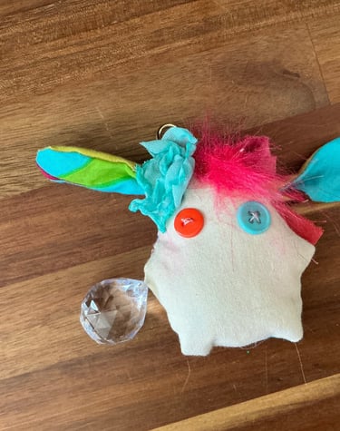 Handmade plush monster keychain with button eyes, colorful fabric ears, and a crystal charm on wood.