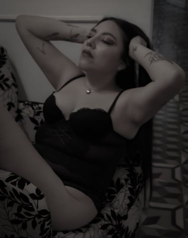 Una mujer con tatuajes en los brazos, vestida con lencería negra de encaje, posando en un sillón