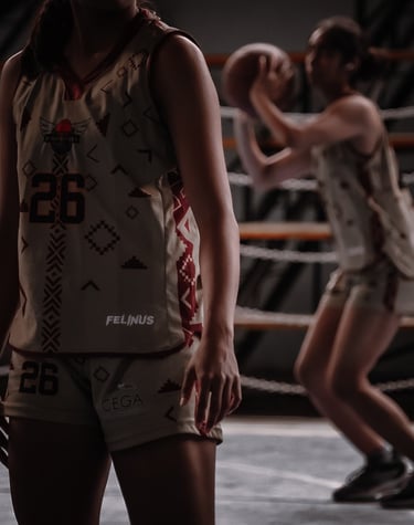 Female athlete wearing a custom patterned basketball jersey while a teammate shoots a ball in the background.