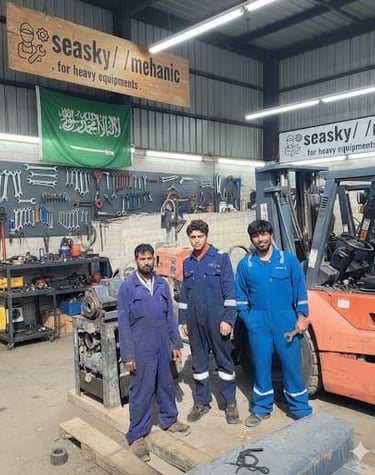 Heavy equipment mechanics in blue coveralls pose in a workshop near a forklift and tool wall.