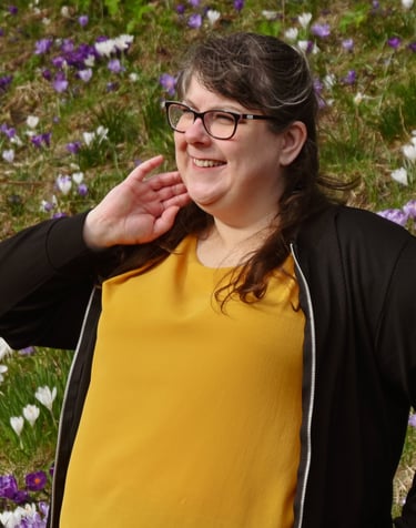 Laura is standing in front of a field of purple and white flowers, looking away from the camera with a soft smile on her face