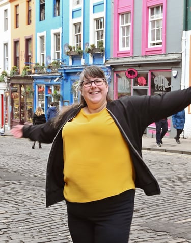 Laura is dancing with her arms out on a cobbled a street with brightly coloured buildings behind her