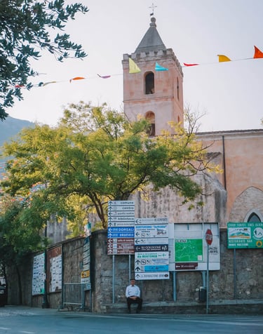 Elderly man in Oliena with street signs and Santa Maria Assunta church tower.