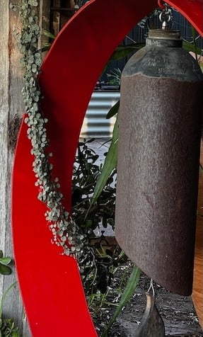 Rustic metal wind chime bell hanging inside a red circular garden sculpture with green vines.