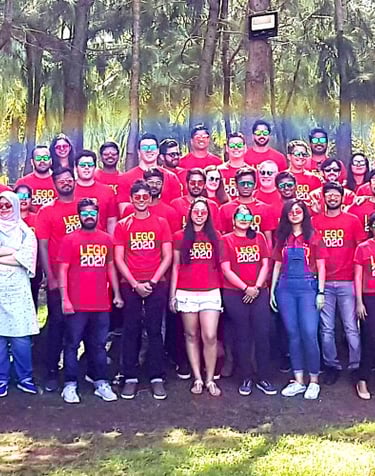 A large group of people wearing red LEGO t-shirts posing for a panoramic photo in a wooded park.