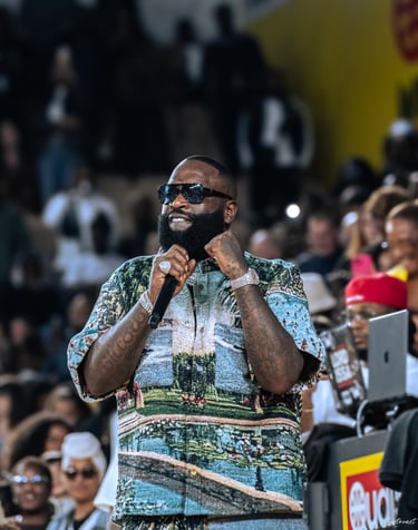 Rapper Rick Ross performing on stage in a scenic tapestry shirt and sunglasses with a microphone.