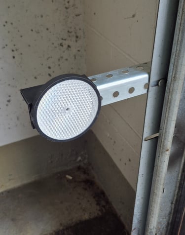 A round white safety reflector mounted on a metal bracket for a garage door sensor system.