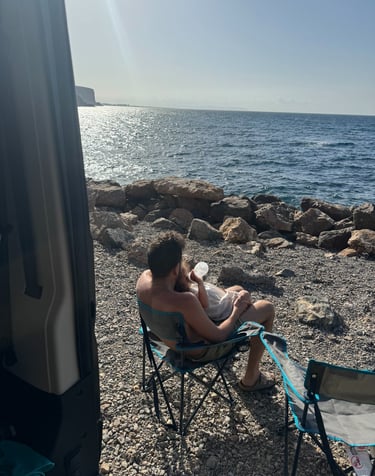 Father feeding a baby on a rocky beach while van life camping by the Mediterranean Sea.