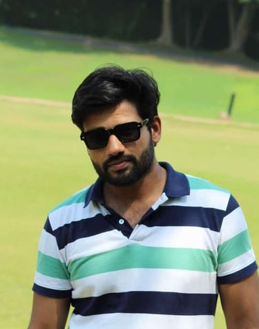 A bearded man wearing black sunglasses and a striped polo shirt posing outdoors in a green field.