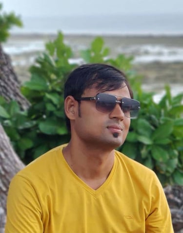 A man wearing a yellow t-shirt and dark sunglasses poses outdoors near lush green foliage and the ocean.