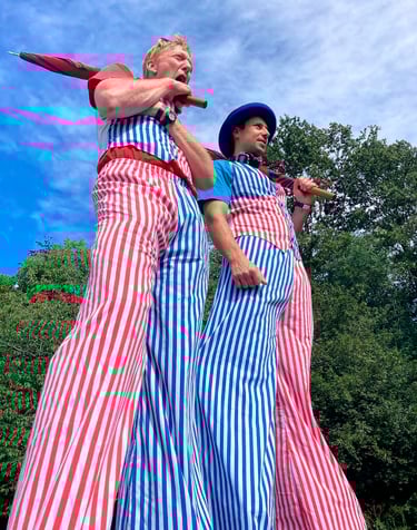 Two circus stilt walkers wearing red and blue striped pants at an outdoor event.