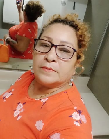 A woman with curly hair and glasses taking a mirror selfie in an orange floral top.
