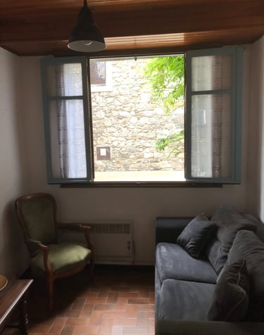 Salon rustique avec canapé gris, fauteuil vert et fenêtre ouverte donnant sur l’église de Quillan