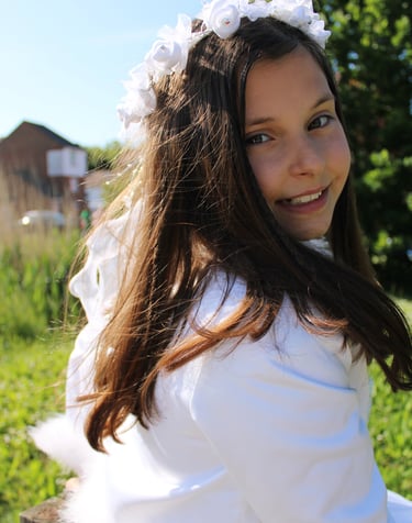 Close-up of a young girl with long brown hair, wearing a white dress and a floral white headband, smiling outdoors on a sunny