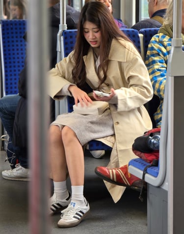 A young woman in a beige coat and white sneakers sits on a tram, looking down into her purse. Other passengers are nearby.