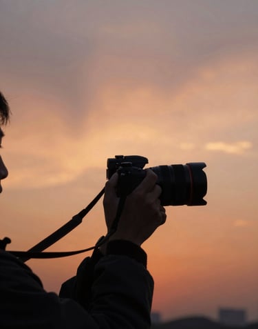 A lifestyle photographer silhouetted against a setting sun, holding a camera in a cinematic, outdoor environment. The sky is filled with warm terracotta and soft sand colors.