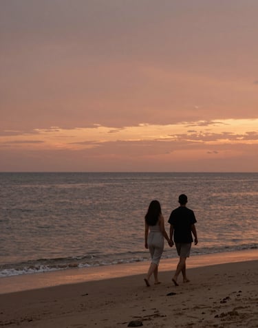 A cinematic shot of a couple walking along a beach at sunset, seen from a distance. The sky is a blend of terracotta and soft sand hues. The water reflects the charcoal tones of the evening light. A sense of movement and authentic storytelling in a sun-drenched environment.