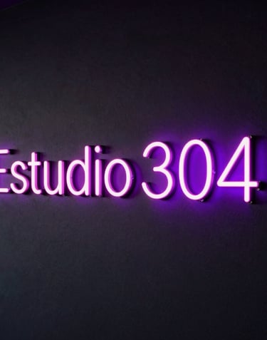 A sleek dark wall featuring the name 'Estudio 304' glowing in vibrant neon pink and neon violet lights, premium modern adult entertainment studio design, dark-mode styling.