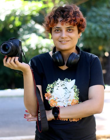 A smiling young female photographer with short curly red hair holds a DSLR camera, wearing headphones around her neck and a