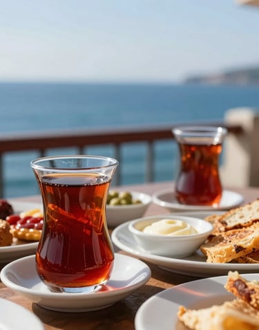 A close-up of a traditional Turkish breakfast spread with a tulip-shaped glass of dark red tea, served on a balcony with the deep blue Black Sea in the background. The lighting is warm and welcoming.