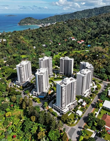 Drone view of a high-end property development area nestled between the hills and the sea in a Southeast Asian / Indonesian island location. Bright midday sun, vibrant greens and deep pine tones.
