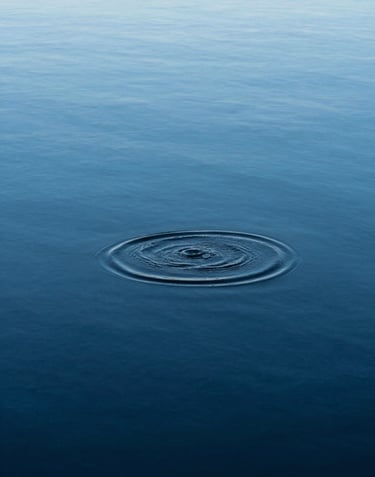 A minimalist, high-angle photograph of a single ripple moving across a perfectly still, deep blue lake. The water is crystalline, reflecting a pale azure sky and absolute stillness.