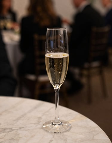 An aesthetic shot of a glass of champagne on a marble table at a luxury event, soft focus background, midnight black and pale beige tones, North American / European gala setting.