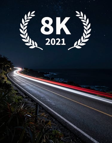 A long-exposure shot of car light trails—red and white—streaking across a dark slate charcoal coastal road under a starry night sky.