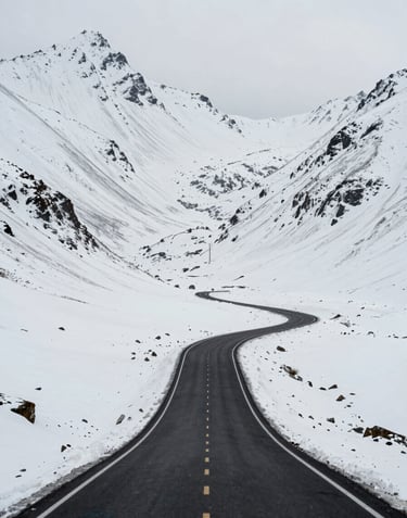 Wide-angle landscape of a high-altitude road cutting through snowy peaks in a Global / International mountain range. Cool off-white snow contrasting with the pitch black asphalt of the road.