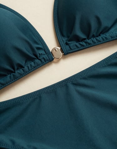 A high-fashion macro shot focusing on the intricate stitching and hardware of a luxury swimsuit. The deep charcoal teal fabric is contrasted by a soft warm cream light, showcasing the premium craftsmanship.