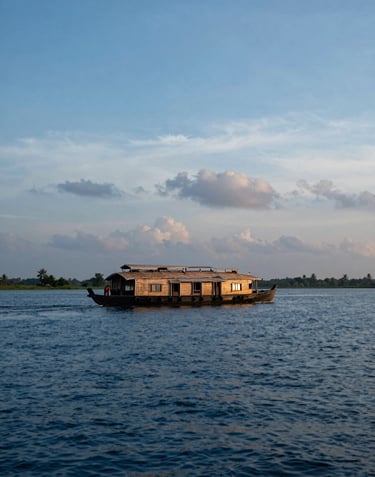 A tranquil view of a houseboat on the Kerala backwaters at sunset. The water is a deep steel blue, and the sky is filled with soft blue and pale grey clouds. Elegant South Asian / Indian landscape.