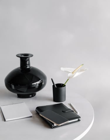 a black vase, a flower in a pencil cup, and notebook on a white desktop