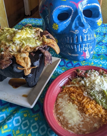 Traditional Mexican molcajete with grilled meats next to rice, beans, and a blue decorative sugar skull.