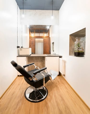 Mały gabinet z czarnym fotelem fryzjerskim, lustrem, umywalką i białymi ścianami.